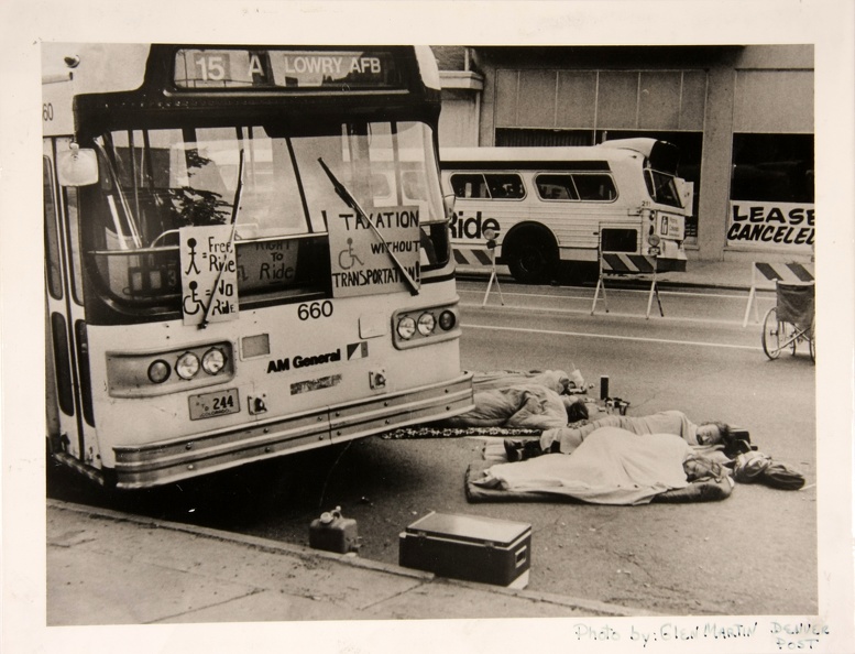 Four men and women are lying wrapped in sleeping bags or blankets on pads in the street in front of a bus. The bus _15 A_ once bound for Lowry AFB_ now appears empty and on the front are 3 handmade posters. Two are outside under the windshield wipers. One says _Taxation without Transportation__ with a drawing of the access symbol_ the other has a picture of a stick figure person next to an equals sign and the words Free Ride_ and then an access symbol guy next to an equals sign and the words No Ride. Inside the window a third sign is partially visible with the access symbol and the words Right to Ride.   There are police_traffic barriers down the middle of the street and a manual wheelchair. There is a bus parked on the opposite side of the street and behind it a city building with a big sign that says _lease canceled._   Around the people lying down are small piles of stuff and there is a cooler by the curb.
